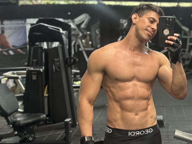 JoseRumBerotop, muscular, shirtless, with defined abs, black underwear, black gloves, short dark hair, holding phone in gym, smiling, looking at reflection, wearing black watch.