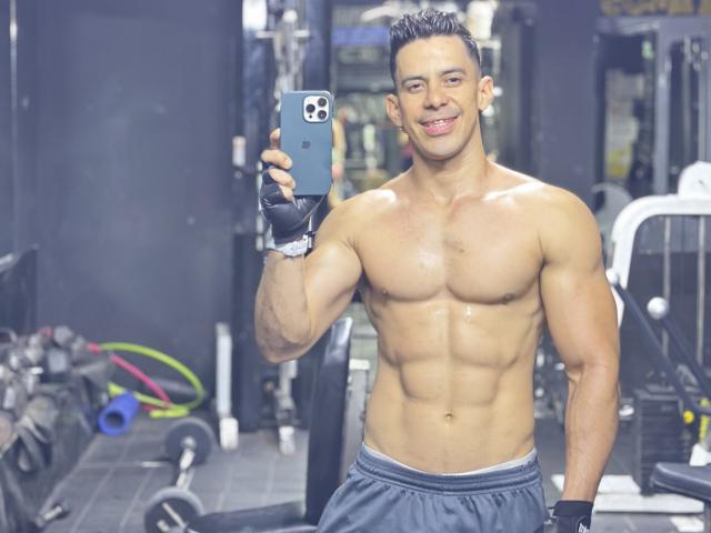 JoseRumBerotop, shirtless with a muscular, shiny, defined ass and chest, holding a smartphone, wearing gym shorts and black fingerless gloves, smiling in a gym.