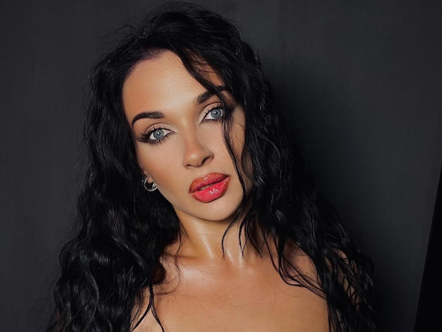 SerenaGlowinski has blue eyes, red glossy lips, and long wavy black hair, with subtle makeup and hoop earrings, looking directly at the camera with a slight, confident smile.