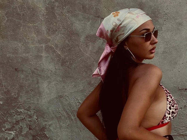 SerenaGlowinski with a leopard print bikini, pink headscarf, and sunglasses, showing her tits and ass, standing against a rough wall, looking back over her shoulder.