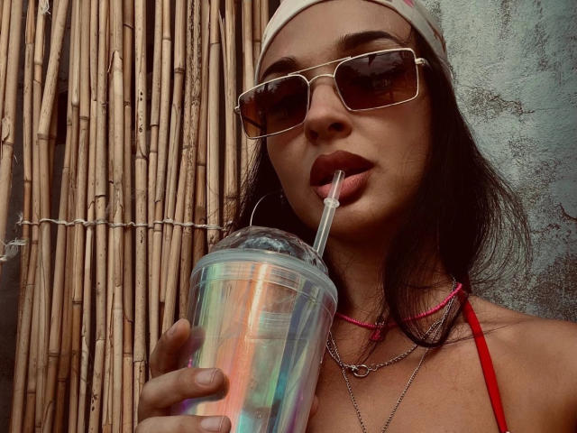 SerenaGlowinski with dark hair, beanie, sunglasses, and red bikini top, drinking from a rainbow iced drink with a straw, wearing pink necklace and silver chain.