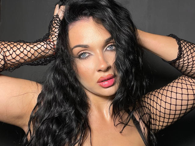 SerenaGlowinski with black fishnet sleeves, hands in wavy black hair, blue eyes, pink lips, and tan skin, wearing a black top, looking seductively at the camera.