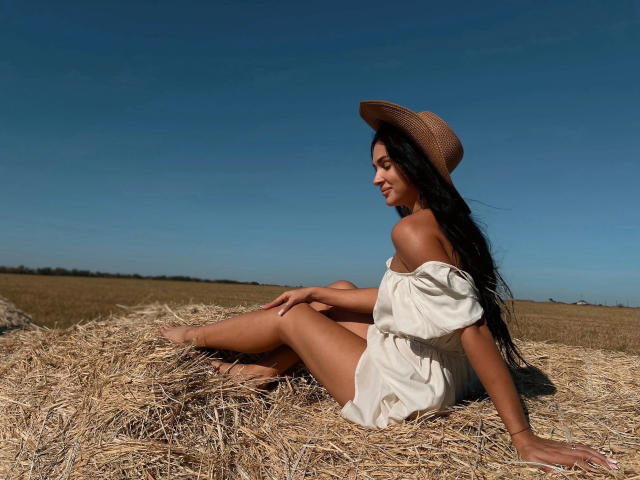SerenaGlowinski with long black hair, tan skin, and a cute smile, sits on a hay bale in a field, wearing a straw hat and off-shoulder white dress.