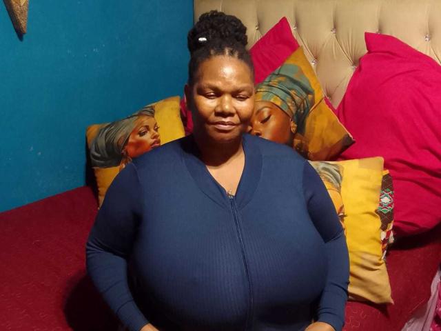 DreamSexyUnique with huge tits, dark skin, and curly hair in a bun, wearing a blue zip-up, sits on a bed with pink and yellow pillows. She's smiling.