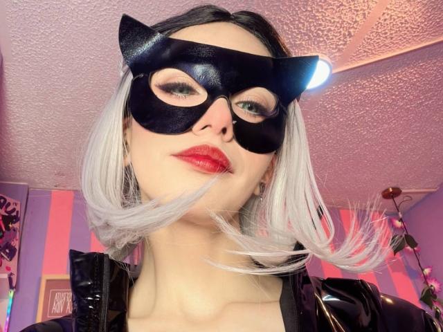 LaurenEvie69 con máscara de gato, lápiz labial rojo, chaqueta de látex negro y cabello corto plateado, mirando hacia abajo, techo blanco, paredes rosas y moradas, ligero reflejo de luz en la máscara, ligero movimiento del cabello por el viento..