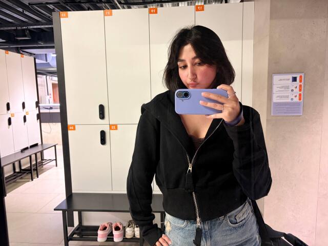 HeikeCarchi with dark hair, pouty lips, wearing a black zip-up hoodie and blue jeans, taking a mirror selfie in a locker room. Her phone is blue. Shoes on bench.