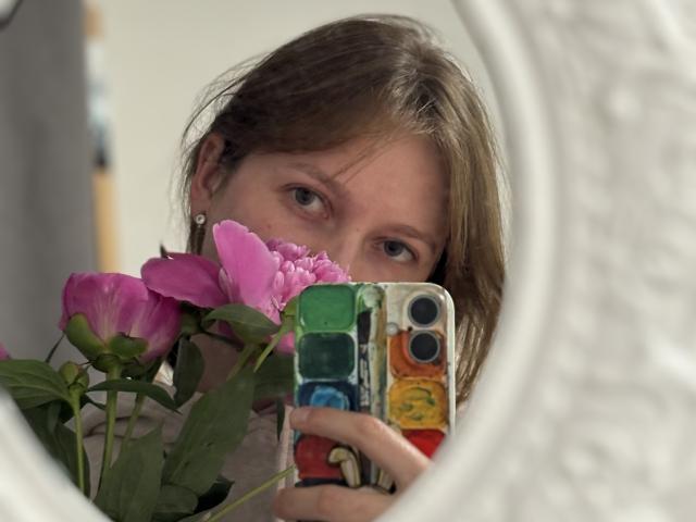 VioletAn with brown hair, blue eyes, holding a phone with colorful buttons, partially hiding behind pink peonies, reflected in a white, textured mirror.