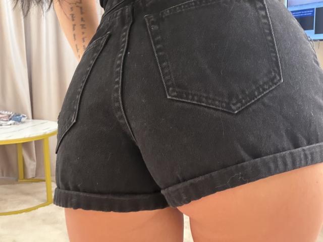 IliriaKara's black denim butt shorts are tight, showing her round ass with two pockets. Her tan skin and tattoo are partially visible. Her shorts are cuffed at the bottom.
