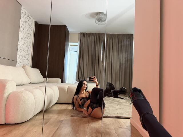 IliriaKara, with long black hair, wearing black lace lingerie, sits on floor, taking selfie in mirror, showing her ass, in modern living room, with white sofa and wooden floor.