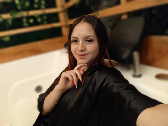 LesleyGray with long red hair, black salon cap, smiling, finger to chin, red nails, white bathtub, wooden wall, holding camera, soft lighting, slight blush, cute.