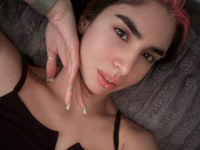TameikaMacclellan, com cabelo com pontas rosa, piercing no nariz e no lábio, vestindo uma blusa preta, com a mão no pescoço, olhos castanhos, unhas longas e maquiagem discreta, olhando para a câmera..