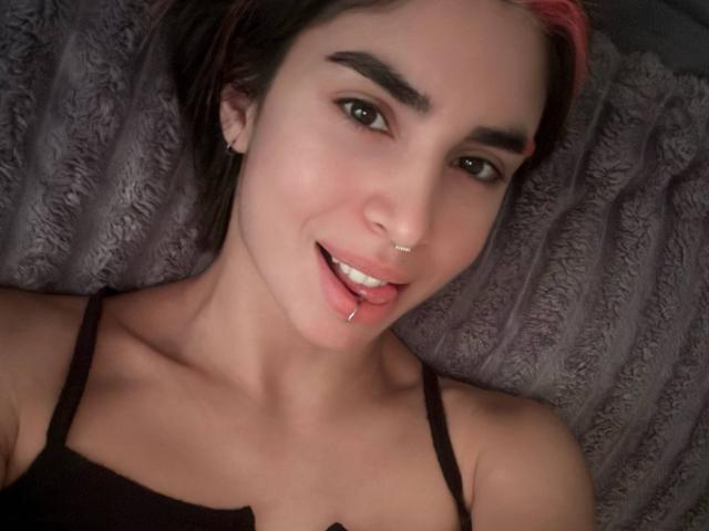 TameikaMacclellan has short brown hair with pink highlights, dark eyebrows, a nose ring, and a tongue piercing, smiling with a black strap top, lying on a gray textured surface.