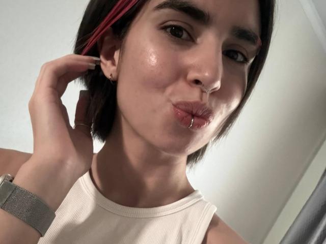 TameikaMacclellan with short black hair and red streak, puckering lips with a nose piercing, wearing a white tank top, and a grey wristwatch, looking at the camera.