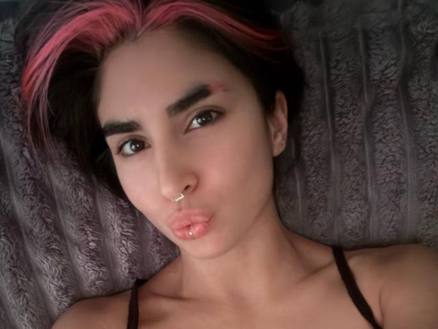 TameikaMacclellan with pink and black hair, nose ring, and pouty lips, lying on a gray textured pillow, wearing a black spaghetti strap top, brown eyes, and light skin.