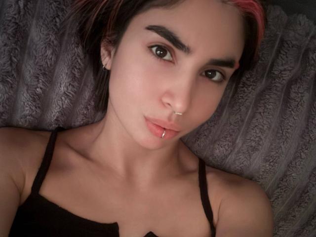 TameikaMacclellan with pink-dyed hair, dark eyebrows, nose ring, lip piercing, black spaghetti straps, lying down on a gray textured pillow, looking at the camera.