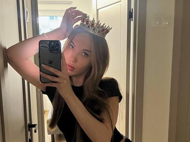 BrettRannels with long brown hair, wearing a black shirt, a silver crown, and a bracelet, taking a mirror selfie with a raised arm, showing off her pouty lips.