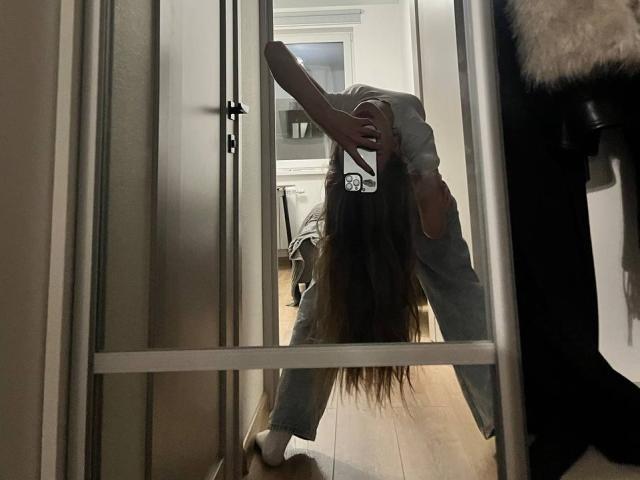BrettRannels is bending over in front of a mirror, with long brown hair hanging down, wearing grey pants and a white shirt, in a dimly lit room.