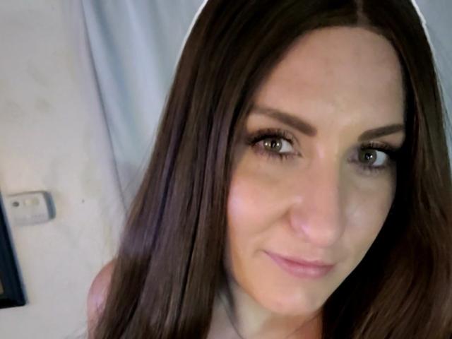 A BackSideBrunette tem cabelos castanhos longos, olhos castanhos e um sorriso discreto, com sobrancelhas visíveis e maquiagem suave, olhando ligeiramente para o lado..