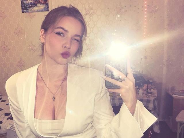 RuthFleetwood with pouty lips, brown hair in a messy bun, wearing a white blazer, exposing her big boobs, taking a mirror selfie with a bright flash.
