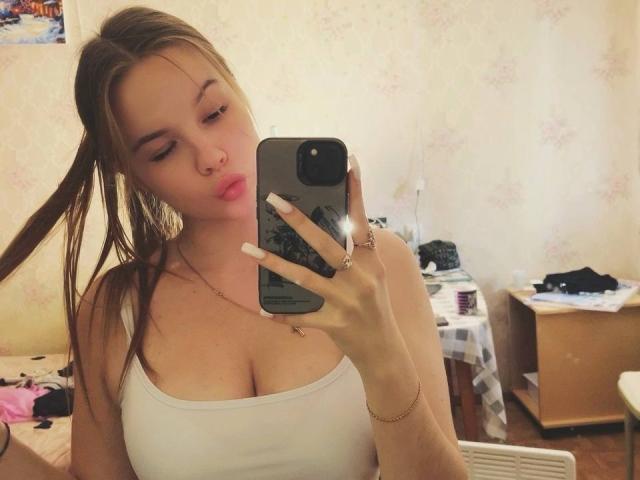RuthFleetwood with pouty lips, brown hair in ponytail, white tank top, showing her tits, holding phone, in messy room, wearing gold bracelet and rings, looking at mirror.