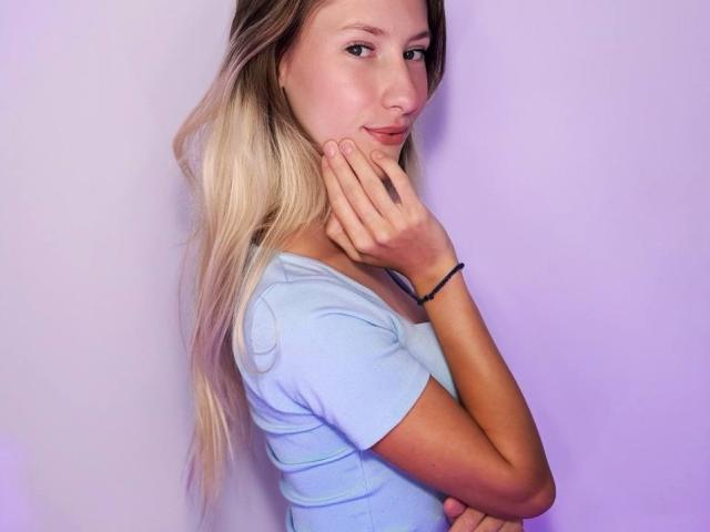 LisaBunnny with long blonde hair, blue shirt, and black bracelet, holding her chin, looking at the camera with a slight smile, hands near her face, soft purple background.