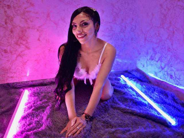 RoxannaMoretti with long black hair, smiling, wearing a pink feathered bra, on all fours on a purple neon lit, fuzzy rug, showing her tits and bare ass.