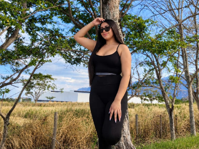 CatalinaRivero stands against a tree, wearing black workout clothes, with long black hair, sunglasses, and a hand touching her head. She has a curvy ass and a small waist.