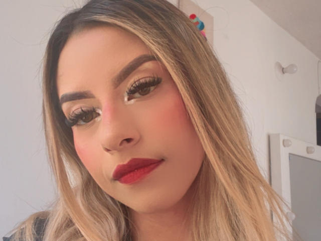 CloeRouss heeft blond haar, rode lippenstift en lange wimpers. Haar wenkbrauwen zijn dik en ze heeft een lichte roze blos op haar wangen. Ze kijkt naar beneden met een subtiele glimlach..