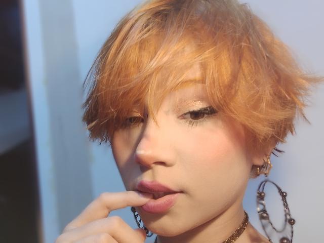ChloeeWilliams with short, messy orange hair, pale skin, and pink lips, biting her finger, wearing a chain necklace and hoop earrings, looking seductive with half-closed eyes.