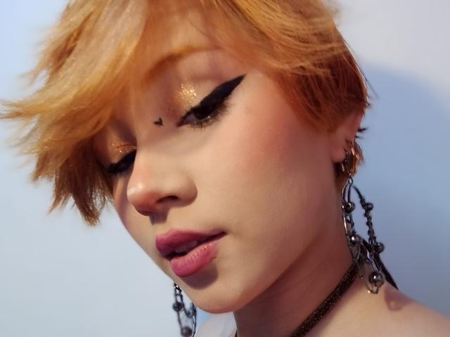 ChloeeWilliams has short orange hair, glittery eye makeup with black winged eyeliner, a small black heart on her forehead, and pink lipstick. She's wearing dangling earrings and a black choker.