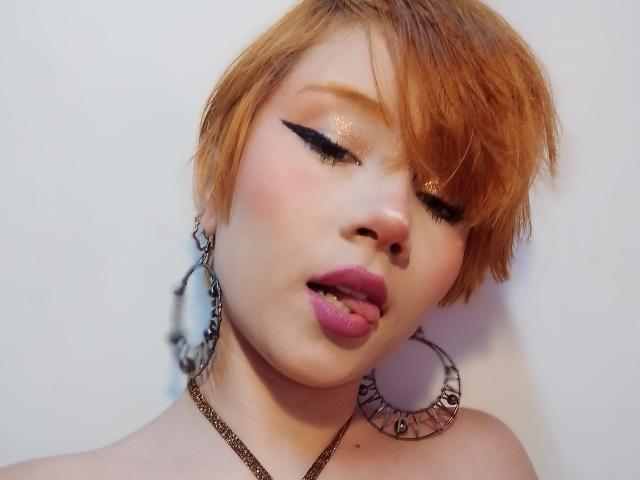 ChloeeWilliams with short orange hair, black eyeliner, shimmery eyeshadow, pink glossy lips, tongue slightly out, wearing large silver hoop earrings, and a brown chain necklace.