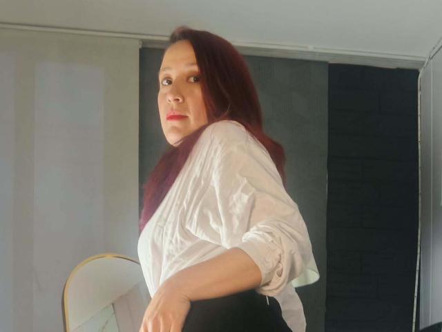 SweettEve with red hair, white shirt, and black skirt, looking over shoulder, hands on waist, standing in front of dark and light curtains, showing her ass.