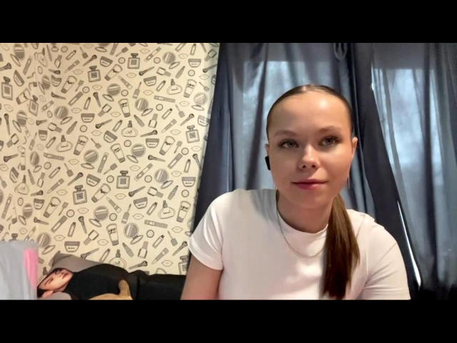 CherrylMeinberg has a slim face with small tits, a white t-shirt, and a ponytail, wearing earbuds, sitting in front of dark curtains, with a patterned wall behind.