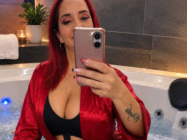 KateMonroe with red hair, wearing a red silky robe, black bra, taking a selfie in a bathroom, showing her large tits and tattoo on her right wrist.