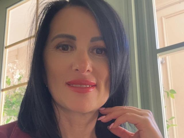 RosaAngie a les cheveux noirs, les yeux marron, du rouge à lèvres, et elle sourit, sa main droite étant près de son menton. Elle porte un haut rouge..