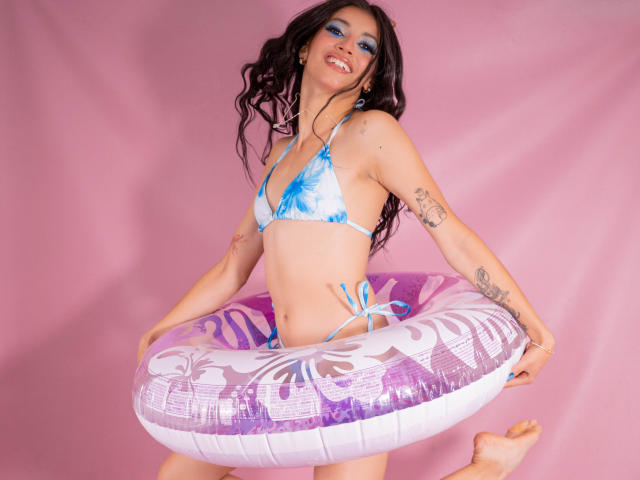 JasminKooper with blue tie-dye bikini, holding purple and white inflatable ring, smiling, tattoos on arms, wavy black hair, pink background, standing, bare feet, confident, sexy.