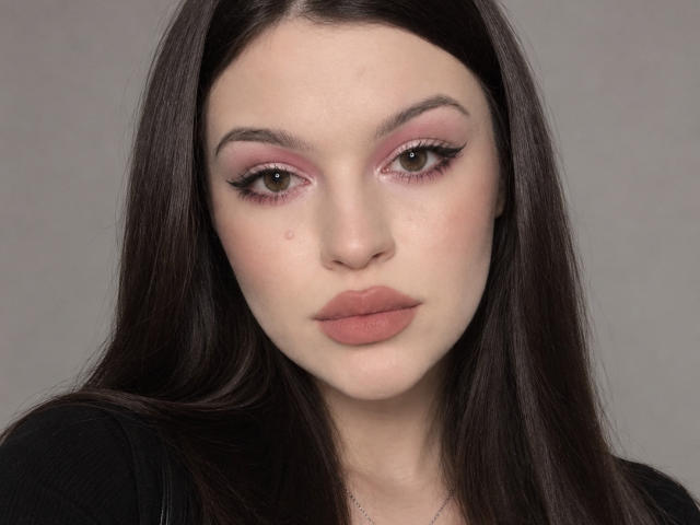 JulietaShy has pale skin, pink eyeshadow, thick black eyeliner, full pink lips, and long dark brown hair, wearing a black top, looking straight into the camera.
