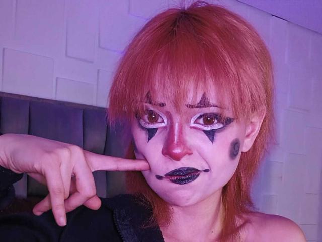 ViolettClay with red hair, white face paint, black star makeup, black lipstick, and purple eyeshadow, pointing at her nose with a finger, wearing a black top.