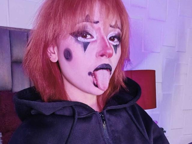 ViolettClay with red hair, black hooded jacket, white face paint with black triangles, and black lipstick, sticking out tongue. Purple and white lighting in the background.