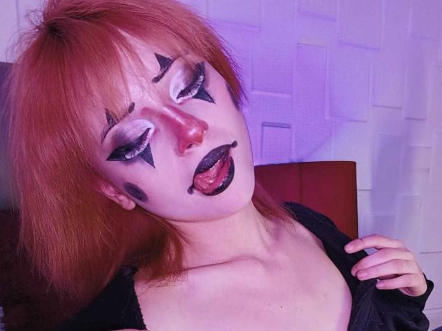 ViolettClay with red hair, white face paint, black star makeup, and black lip paint, tongue out, wearing a black top, hand on chest.