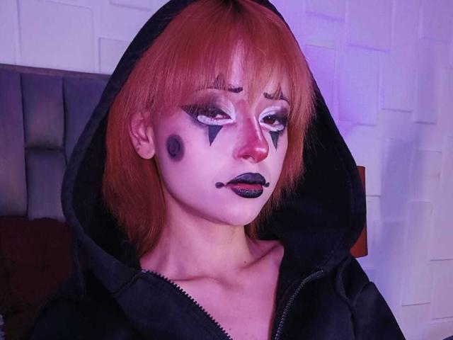 ViolettClay with orange hair, black hood, white face paint, dark eyeliner, black lipstick, and black makeup marks, wearing a black hoodie, looking serious.