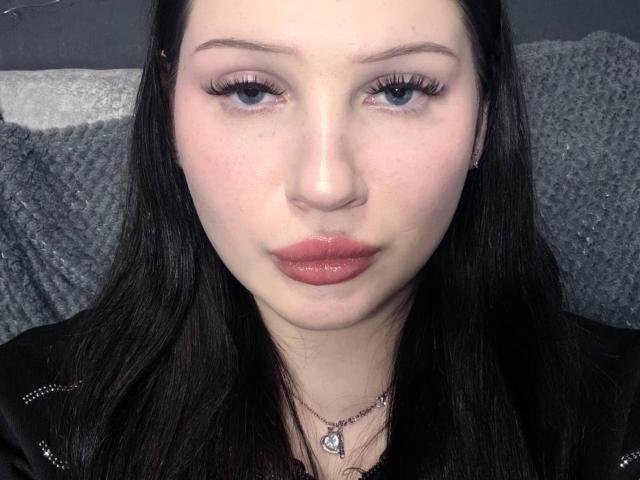 StasiaFreshley has pale skin, dark straight hair, blue eyes, and glossy pink lips. She's wearing a black jacket with a heart necklace, looking directly at the camera.