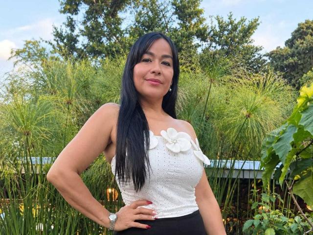 TamaraFoxy has black hair, medium tits, and a cute smile. She's wearing a white floral top, black skirt, and red nail polish, standing in a green garden.