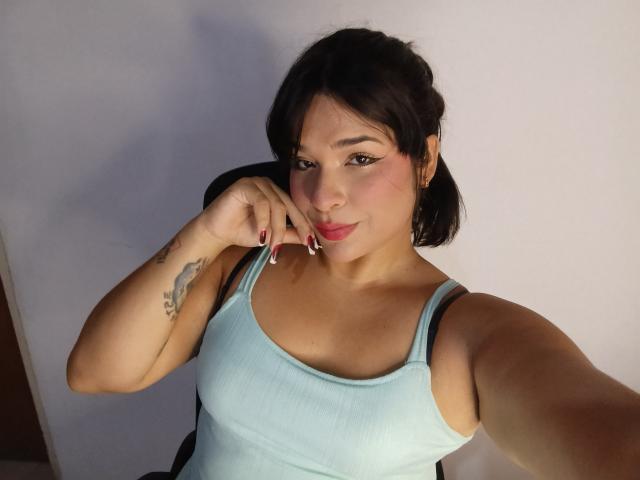 AntonelaPark with short black hair, light blue tank top, small tits, tattoo on left arm, holding finger to lips, pink lipstick, light skin, sitting in chair, selfie.