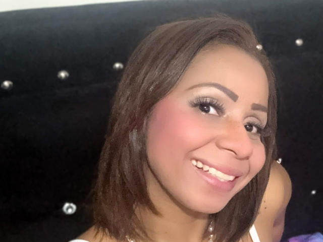 AlondraMonique has brown skin, medium-length brown hair, pink blush, and white makeup with long eyelashes. She's smiling with her nose slightly up, showing her teeth.