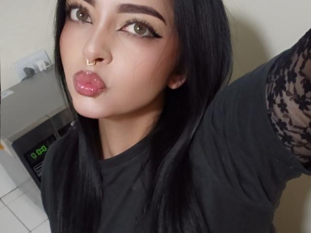 LilithAmoth, con i capelli neri, un anello al naso e labbra rosa lucide, che fa la smorfia, indossa una maglietta nera con maniche in pizzo, in un selfie. I suoi occhi verdi sono leggermente rivolti verso l'alto..