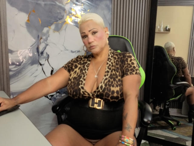 Gianella Cox tiene senos grandes, un trasero regordete y cabello rubio corto. Lleva una blusa de estampado de leopardo, un cinturón negro y está sentada en una silla verde, luciendo seria con una expresión neutra..
