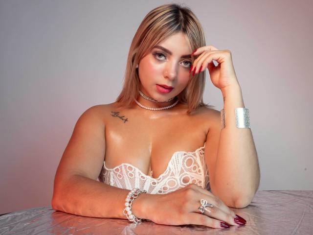 CloeEvanss with blonde hair, wearing white lace bodice, shiny skin, red nails, choker, bracelet, ring, and tattoo, looking at camera with hand on head. Boobs, ass visible.