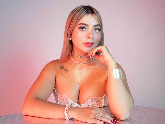 CloeEvanss with blonde hair, pink lipstick, white lace bra, pearl necklace, silver bracelet, and tattoo, resting chin on hand, showing her tits and red nails.