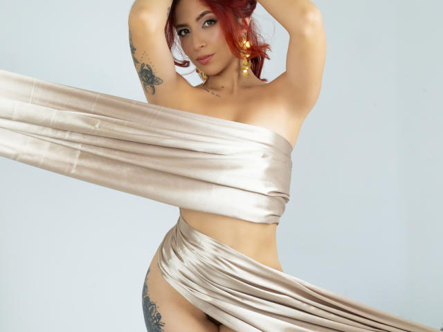 MiaaRouge with red hair, gold earrings, tattoos, and a shiny beige wrap covering her tits and ass, arms raised, looking at the camera, in a studio shot.